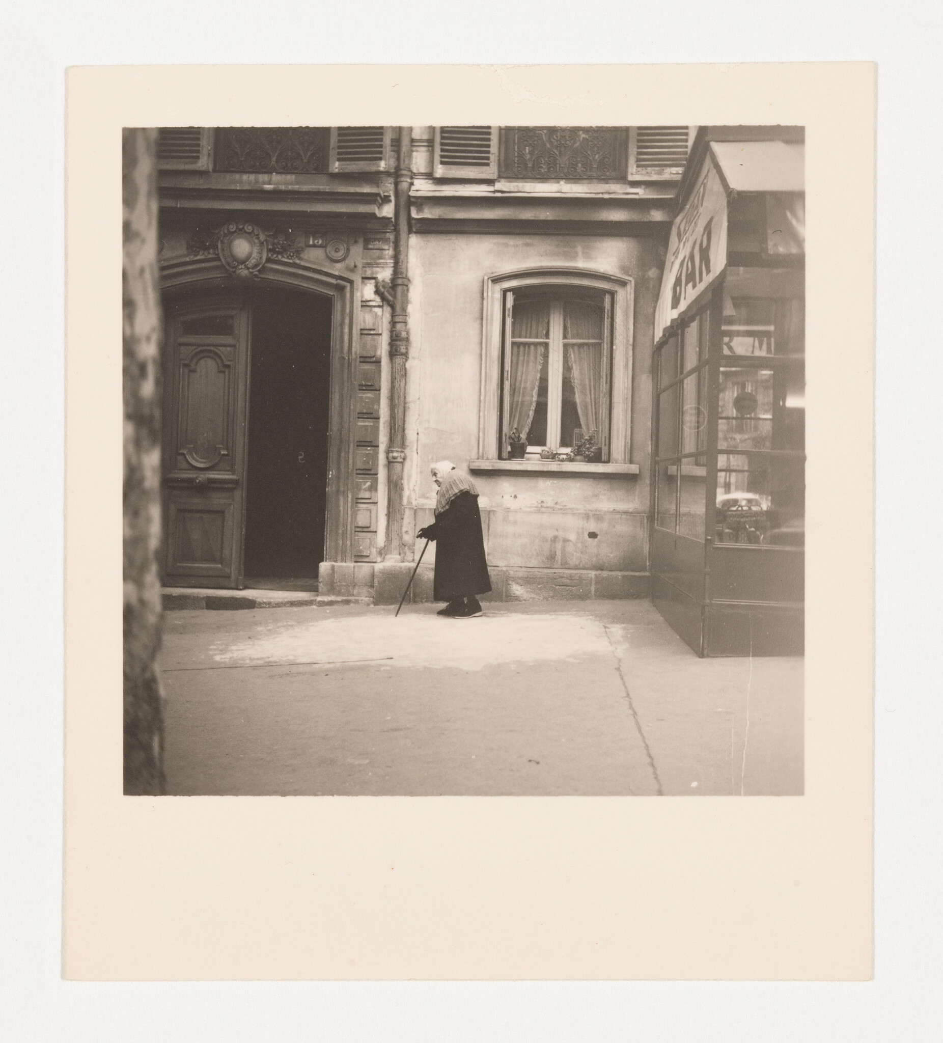 An elderly woman with a cane walks slowly past a building entrance and shop window.