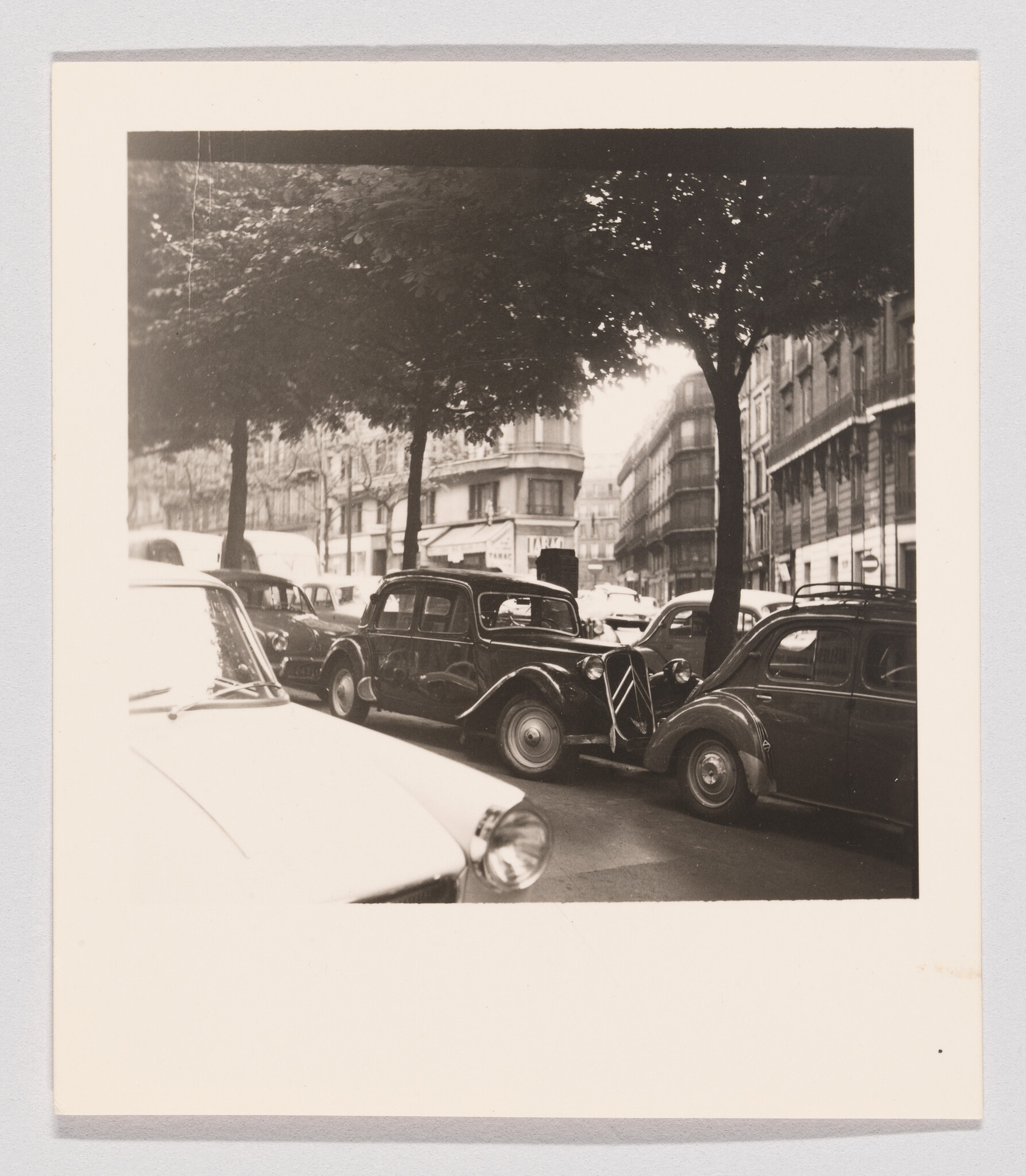 A row of vintage cars parked under trees along a busy city street with buildings.