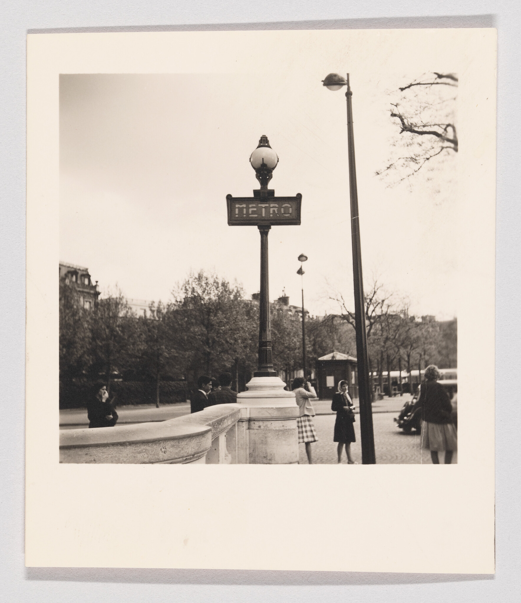 A vintage Metro sign atop a lamp post with pedestrians standing nearby on the sidewalk.