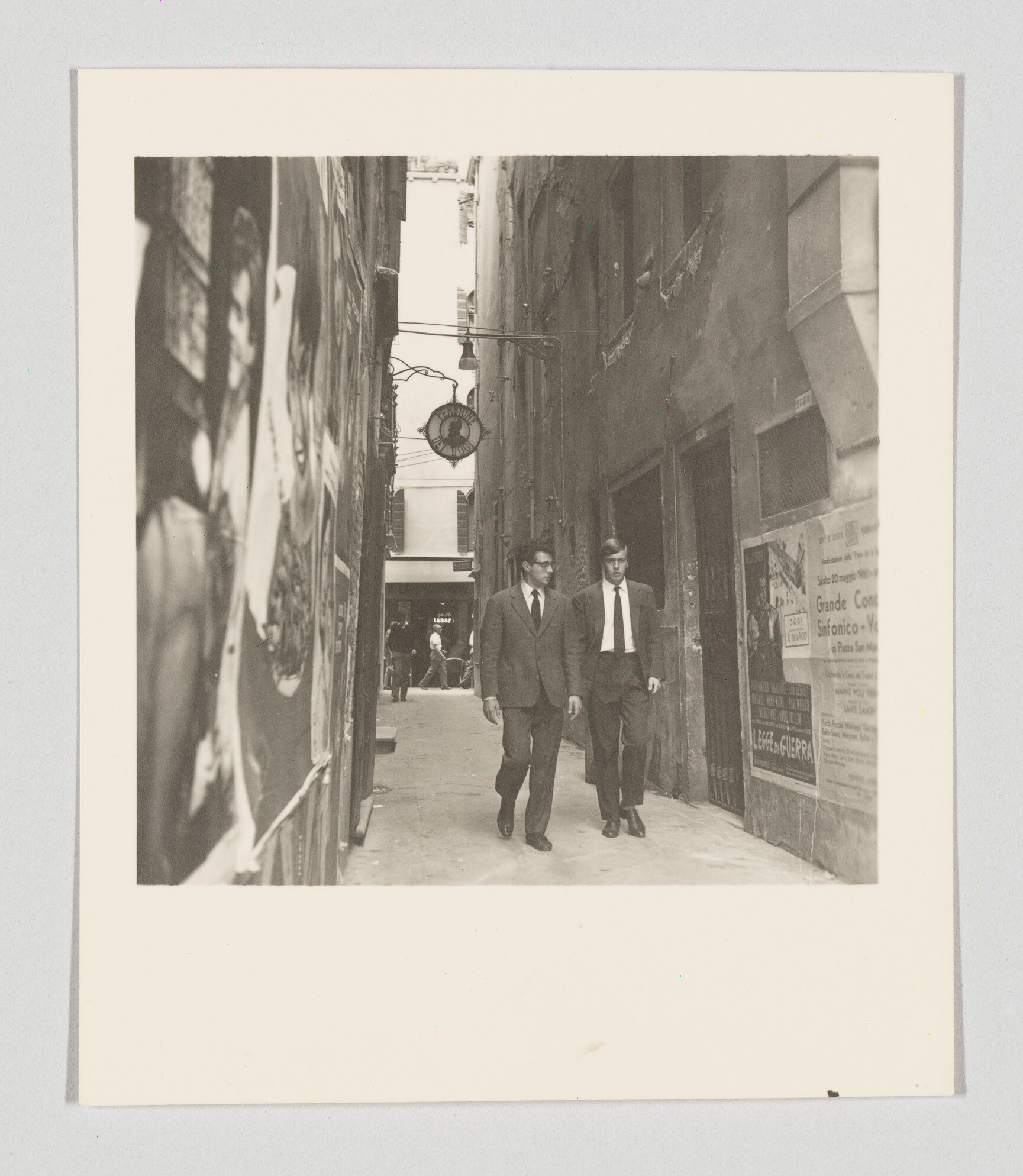 Two men in suits walk down a narrow alley lined with posters and old buildings.