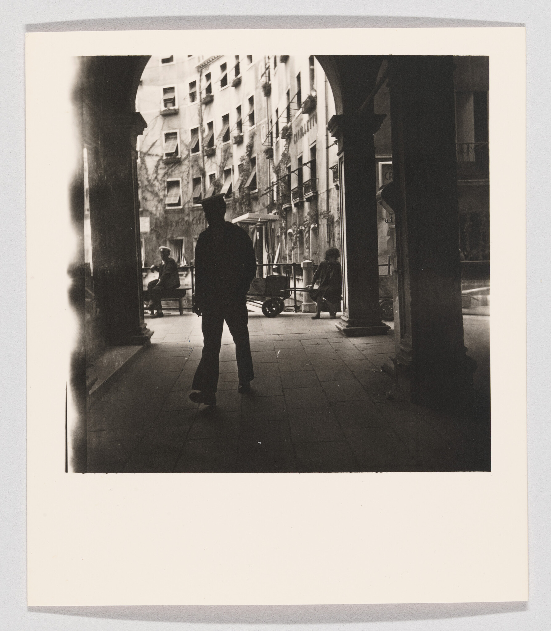 A black and white photograph depicting a silhouetted figure standing in a sunlit arcade with classical columns. The background shows a street scene with buildings, a few pedestrians, and a parked bicycle. The contrast between light and shadow creates a dramatic atmosphere.