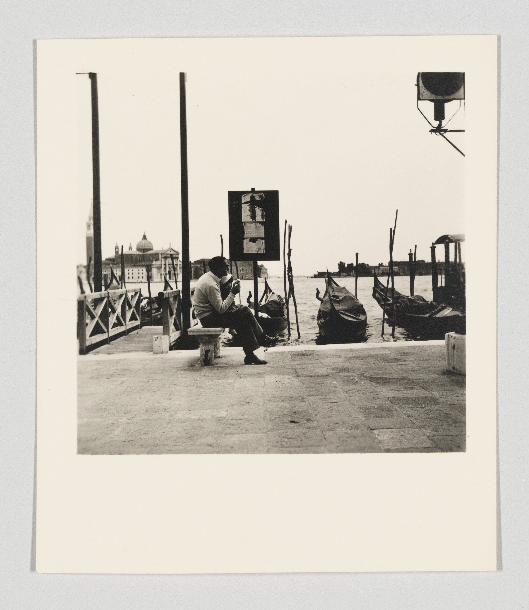 A man sits on a bench by the water eating a sandwich as gondolas wait.