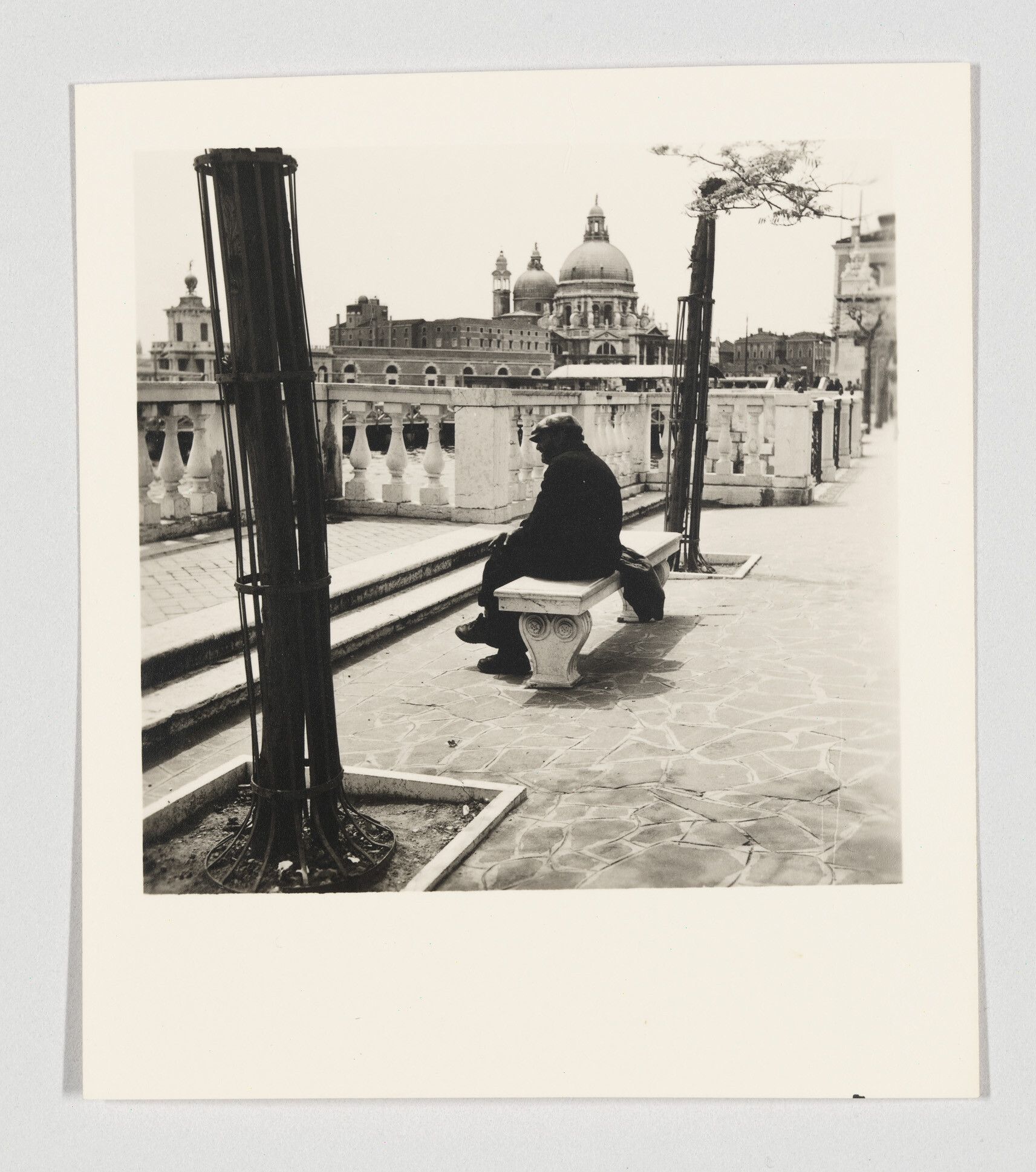 A solitary person sits on a bench by a riverside balustrade with a domed cathedral behind.
