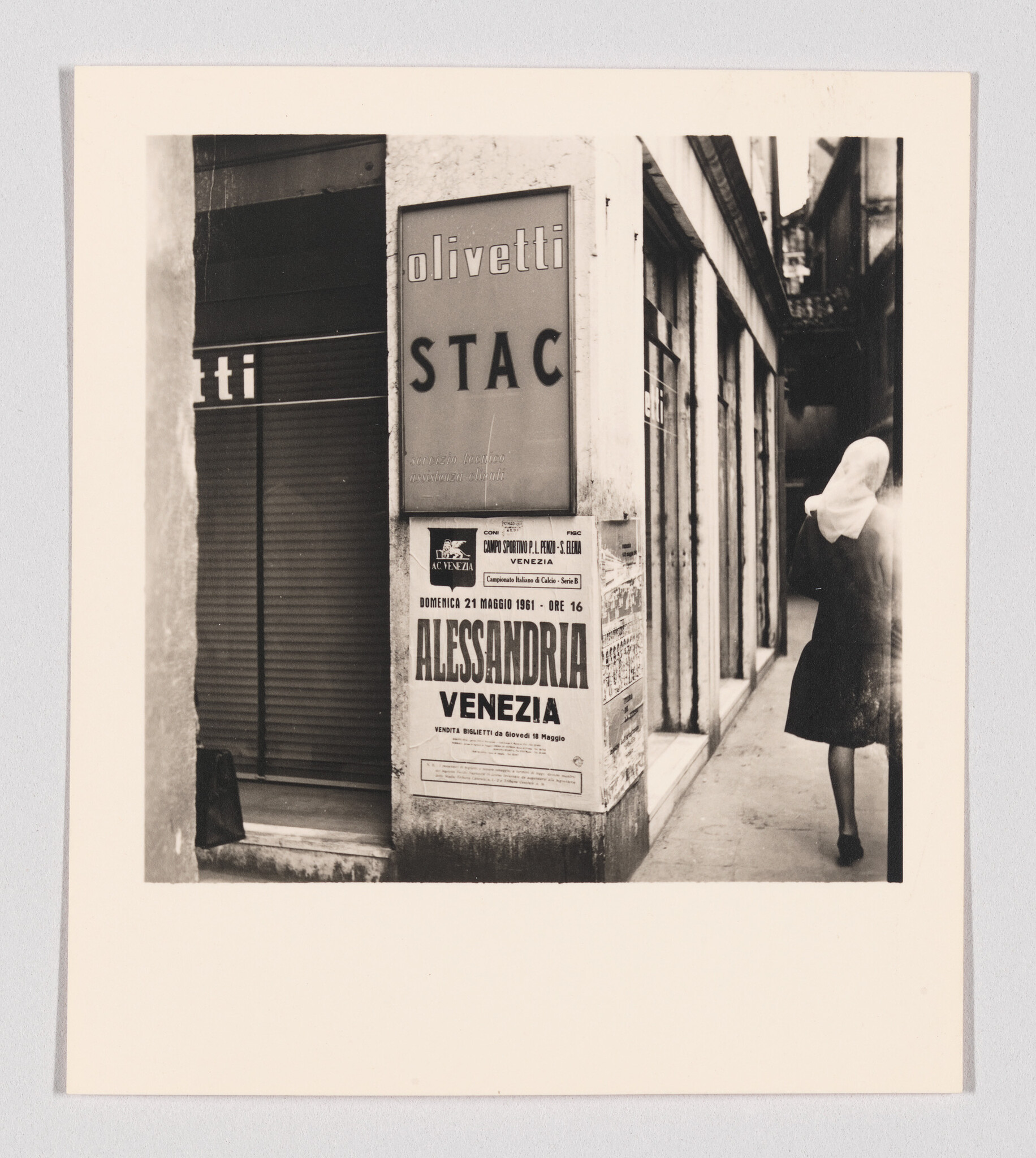 Woman wearing a headscarf walks down a narrow alley past an Alessandria Venezia poster.