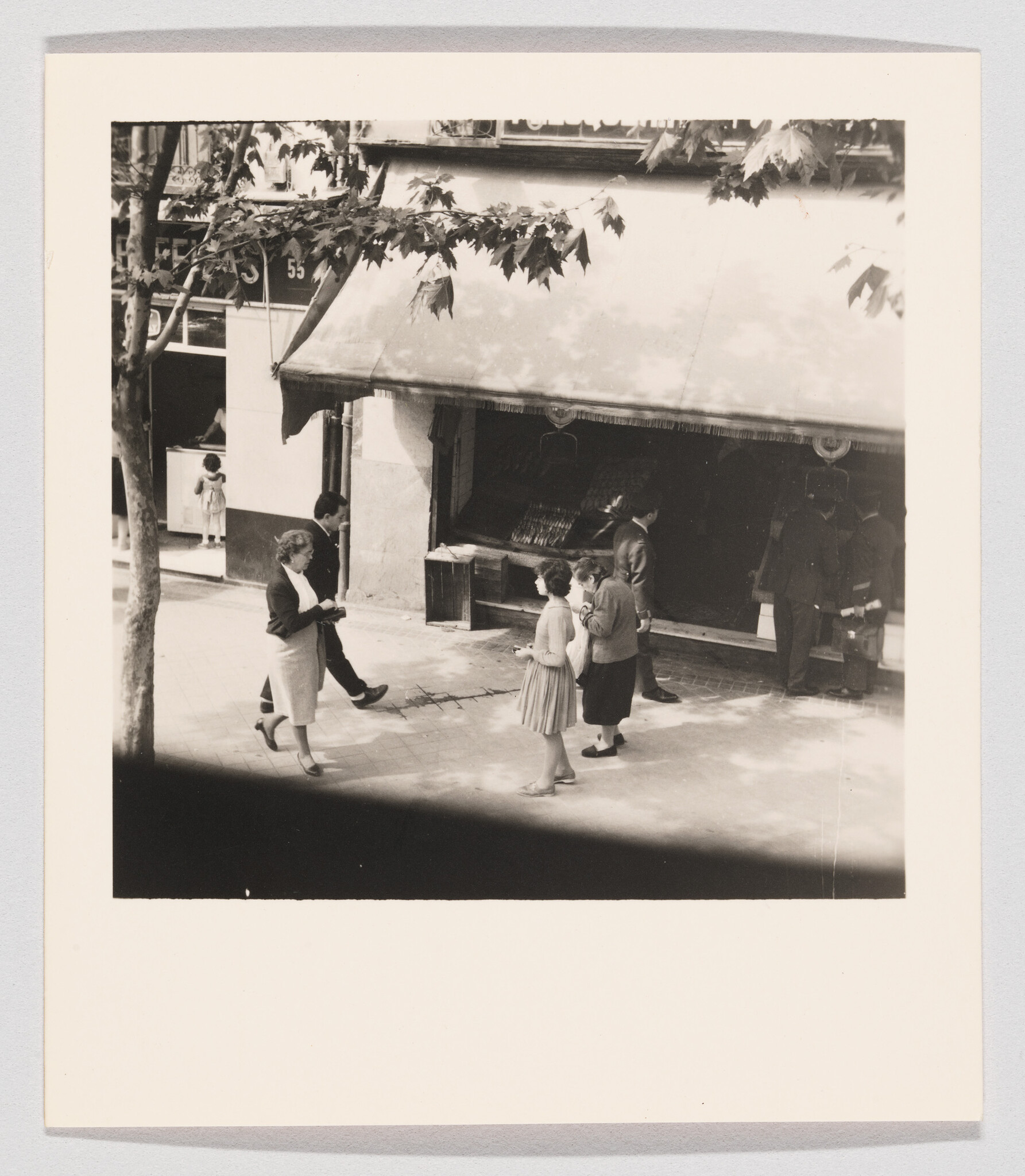 A small group of people stand and walk outside a shop under a large awning on the sidewalk.