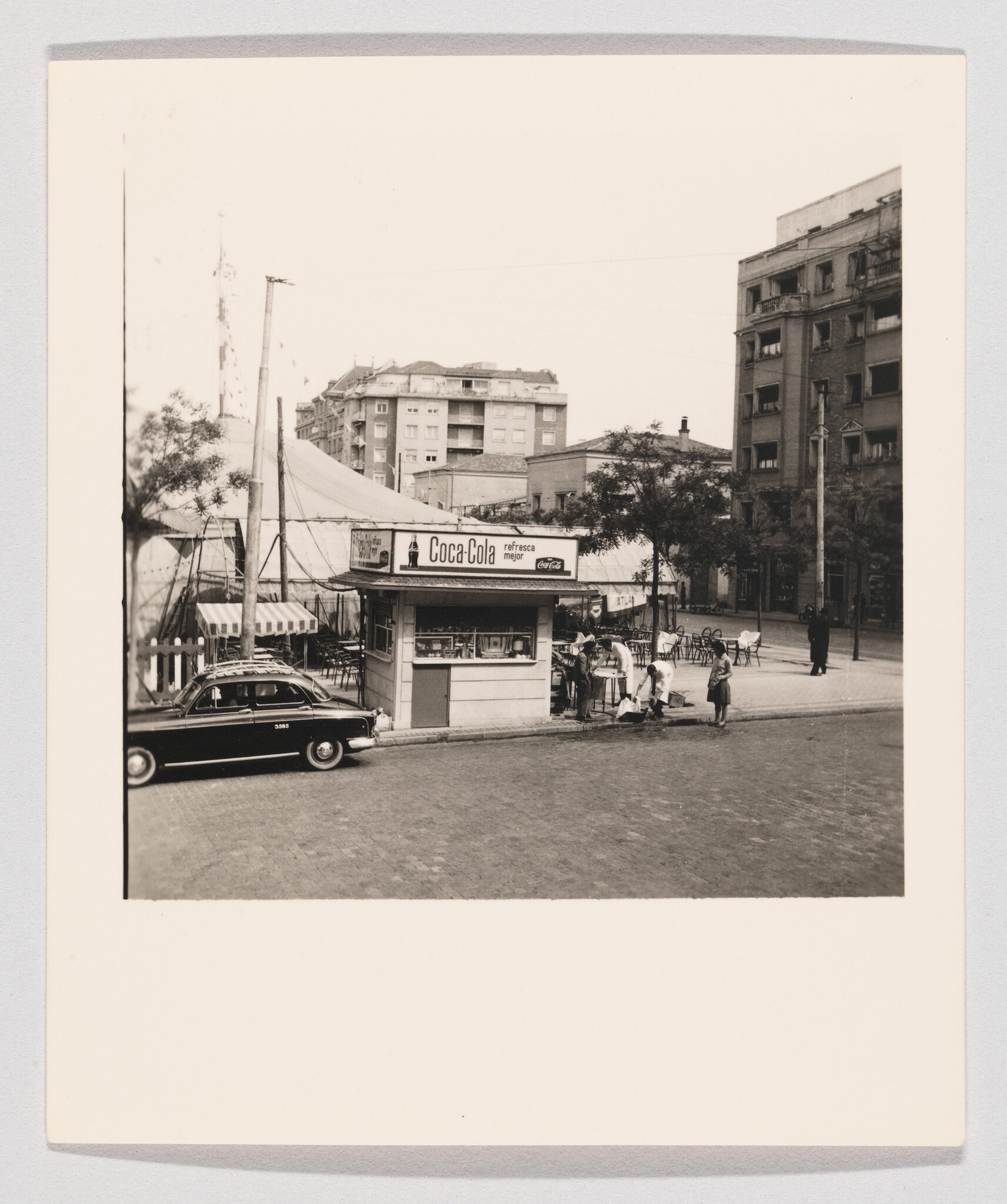 A small Coca-Cola kiosk on a city street with people gathered and a parked car.