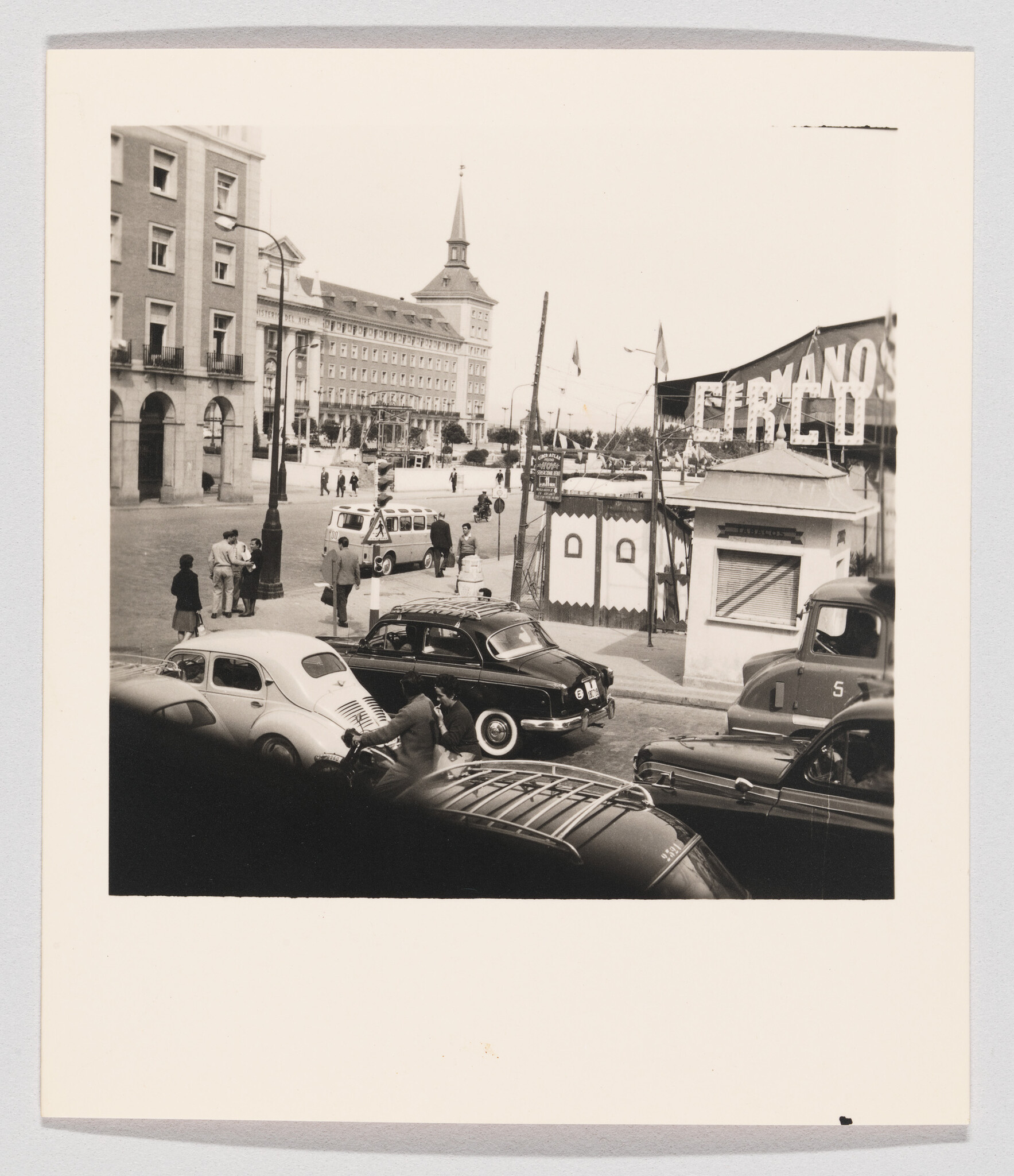 Vintage cars and people gathered near a circus entrance in a city square.