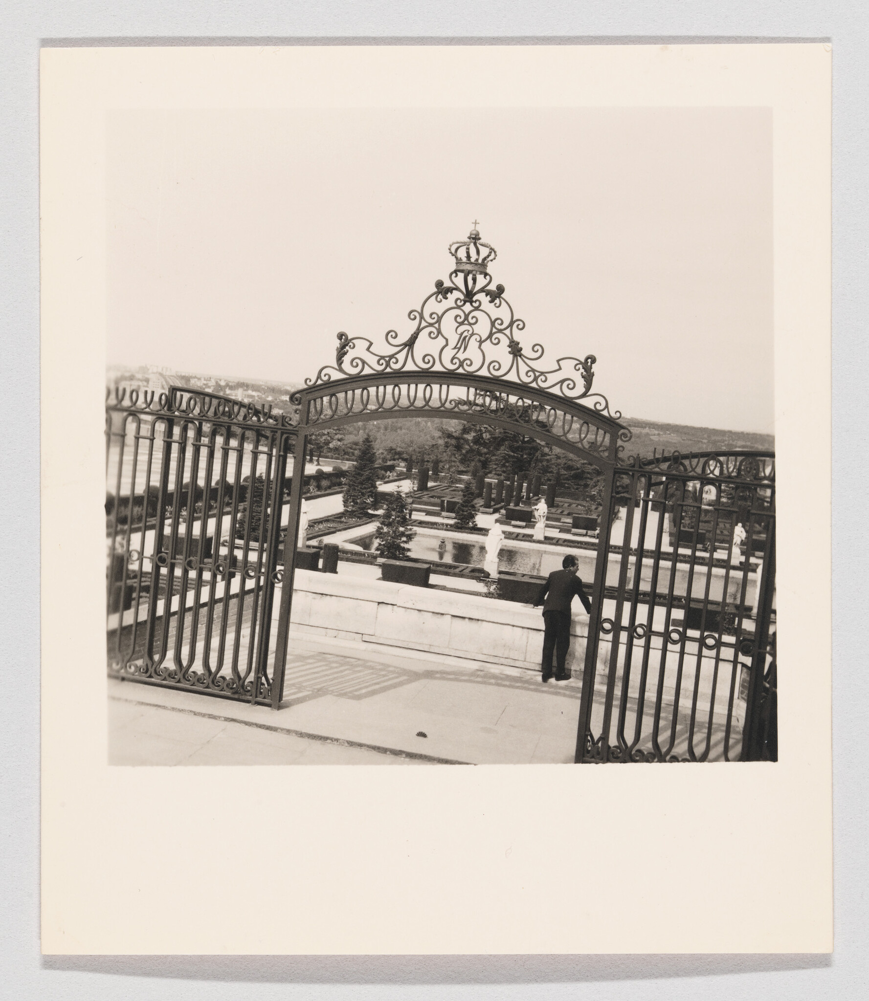 Ornate wrought-iron gates open onto a formal garden while a person leans on the railing.