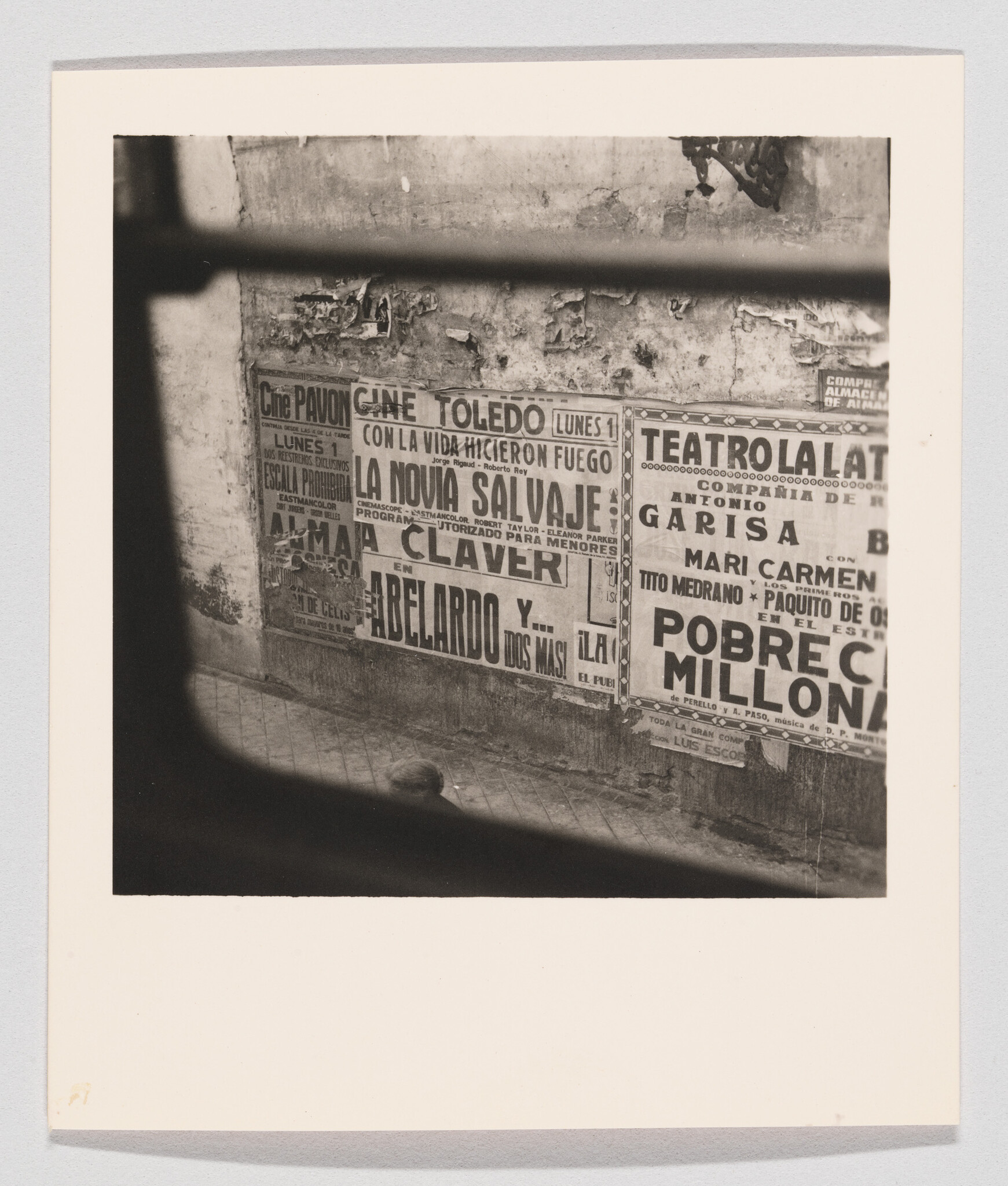 Weathered wall plastered with vintage Spanish theater and film posters seen through window bars.
