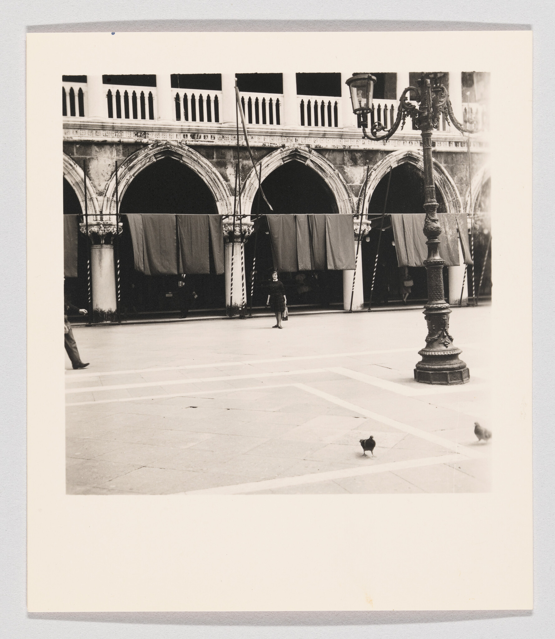 A black and white photograph depicting a spacious square with patterned paving stones. In the background, there's an elegant building with arched openings and curtains, possibly a historic arcade or gallery. A solitary figure walks across the square, and a classic street lamp stands prominently in the foreground. A couple of pigeons are visible on the ground, adding life to the serene scene. The photo has a white border, suggesting it may be a print or part of an album.