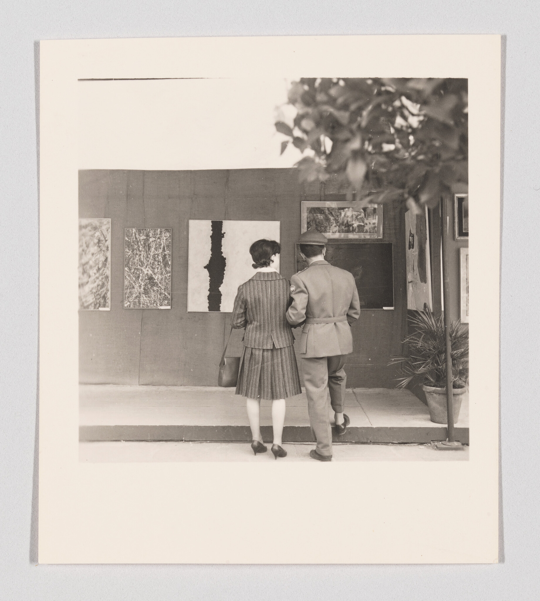 A woman and a uniformed man stand arm-in-arm looking at paintings displayed on a gallery wall.
