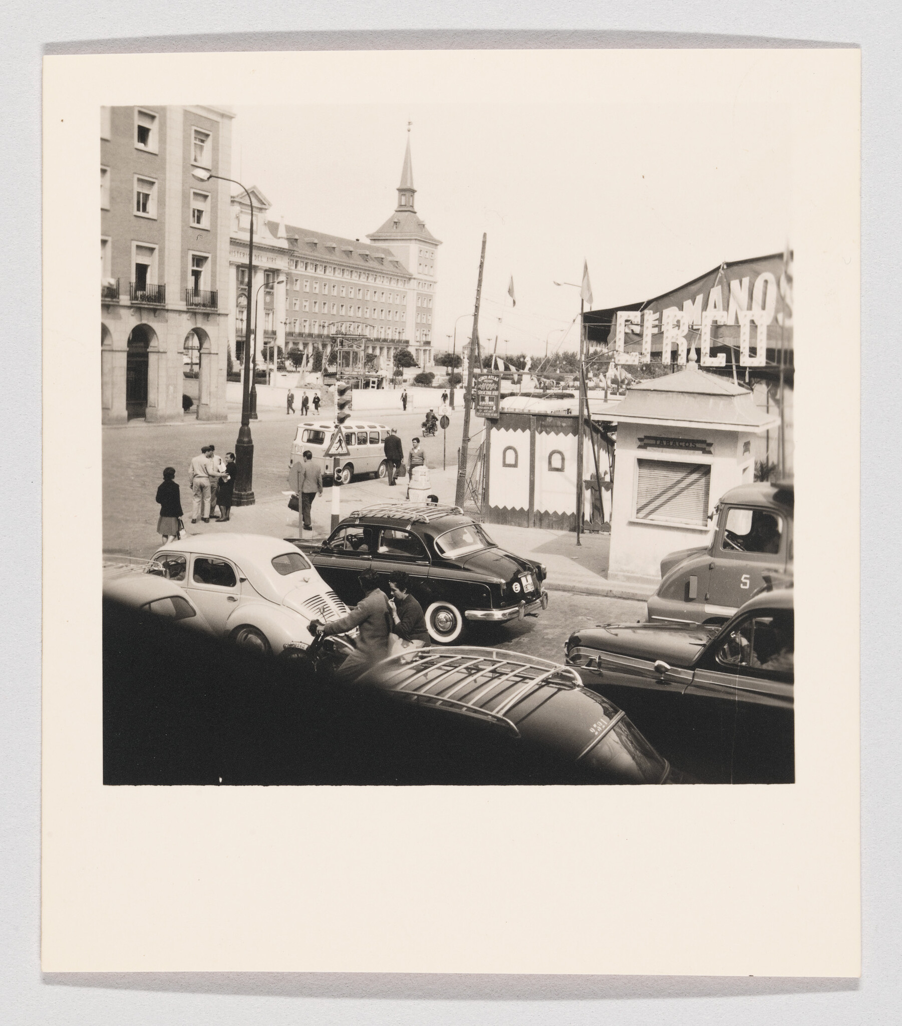 Several vintage cars and people gather on a busy city street near a small fair kiosk.