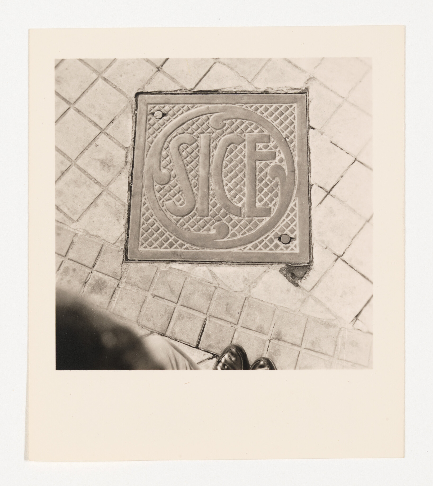 Square manhole cover with circular pattern set in tiled sidewalk with visible shoes.