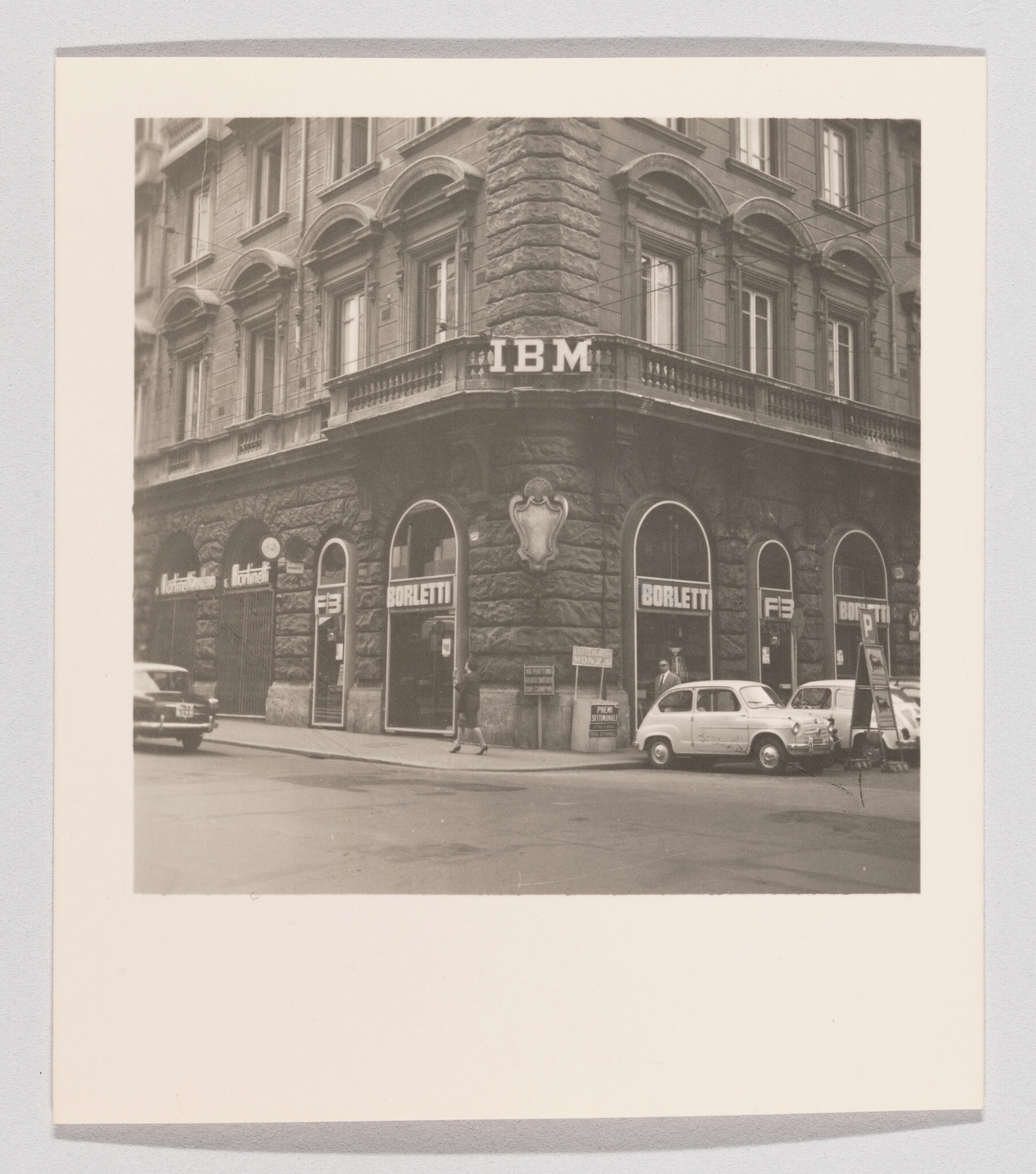 Corner stone building with IBM sign and Borletti storefronts while people walk and cars park.