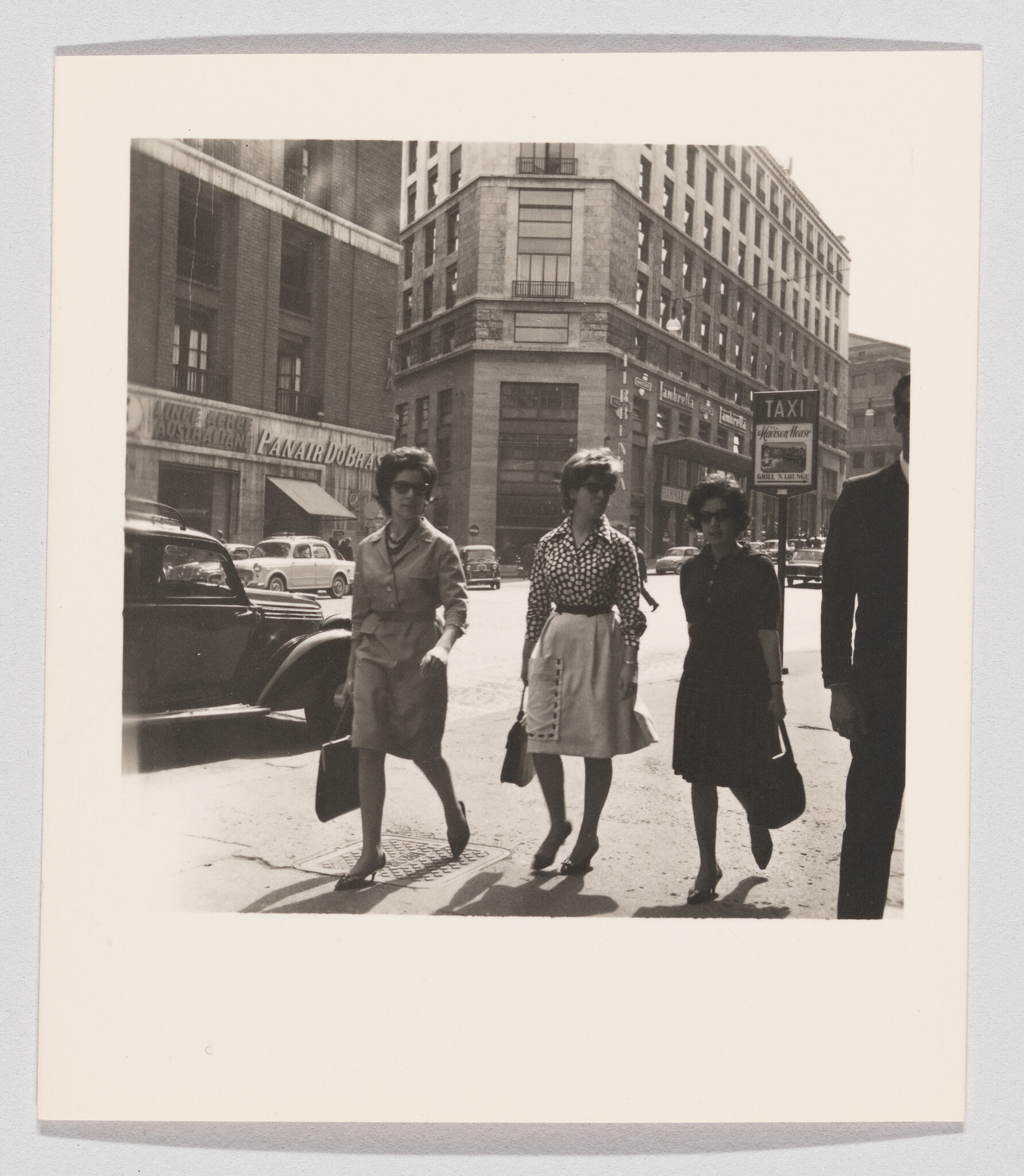 Three women walk across a busy city street carrying purses with cars and tall buildings behind them.