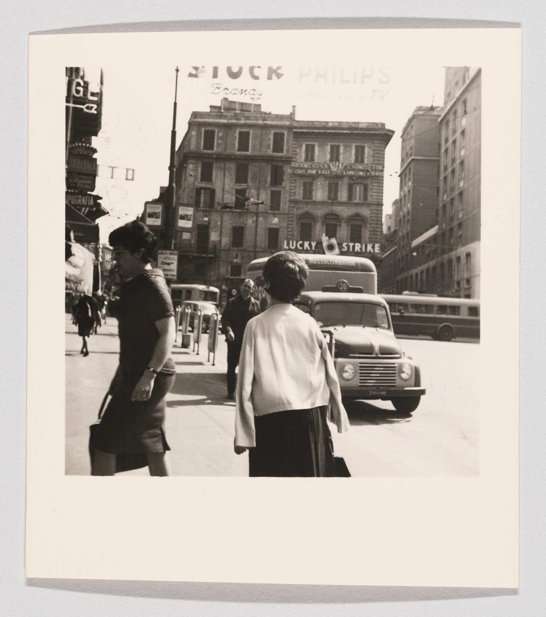 Two women walk along a busy city street with a parked van and a lucky strike sign overhead.