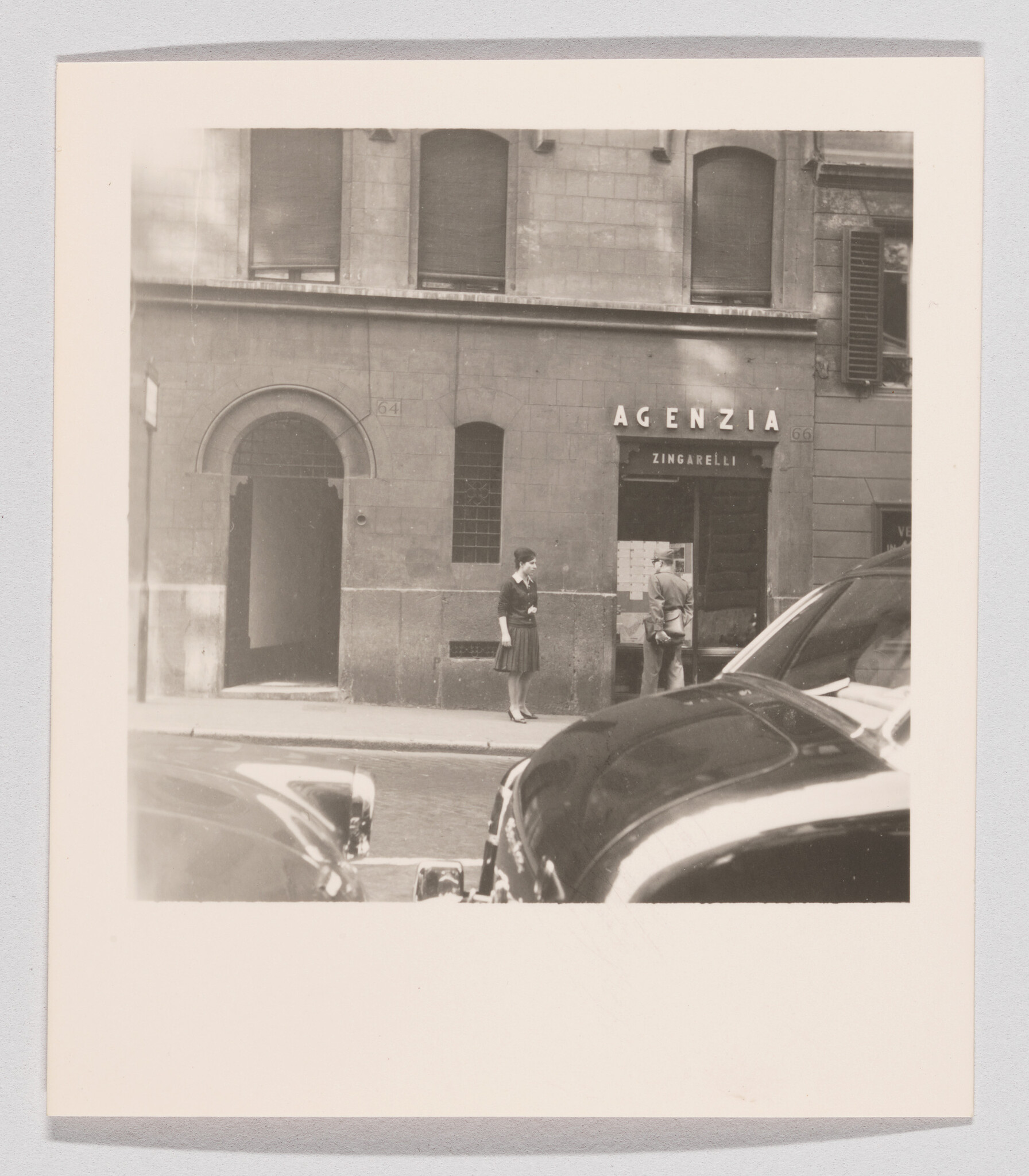 A woman and man stand outside the Agenzia Zingarelli office while cars park in the foreground.
