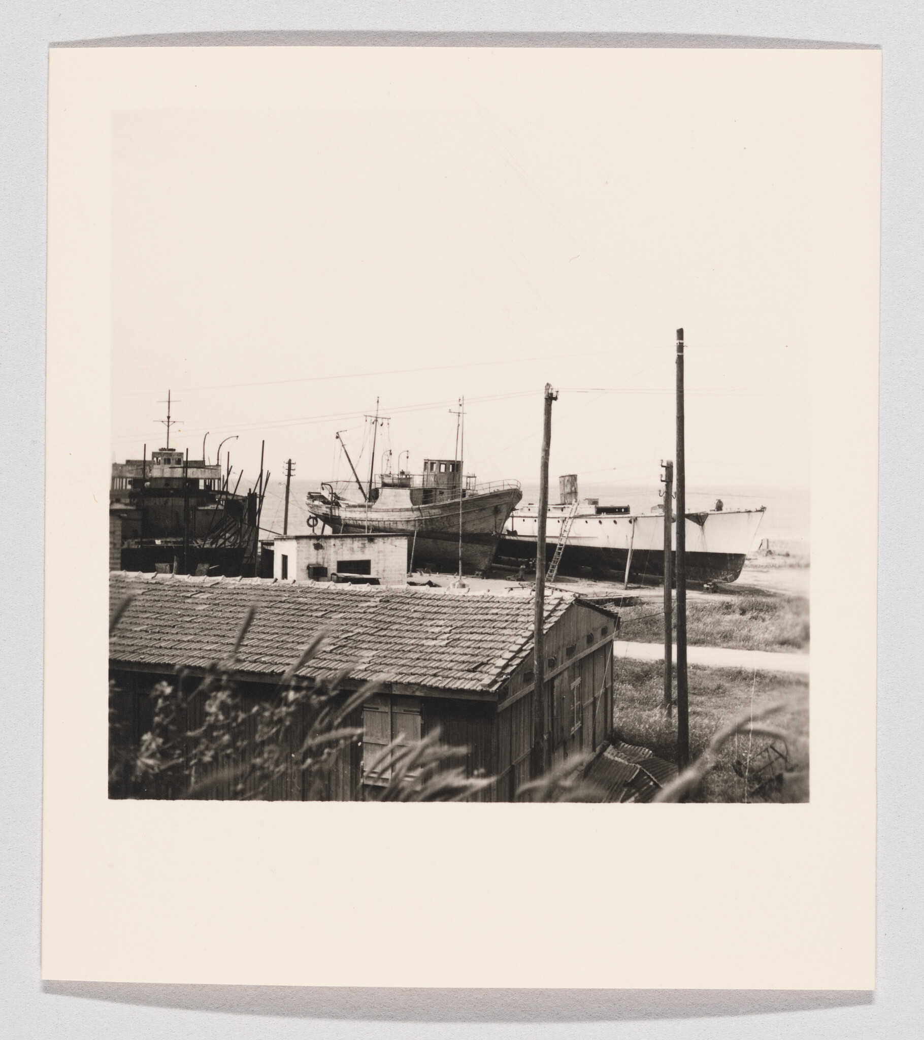 Several fishing boats sit on the shore behind a corrugated-roof shed with utility poles.