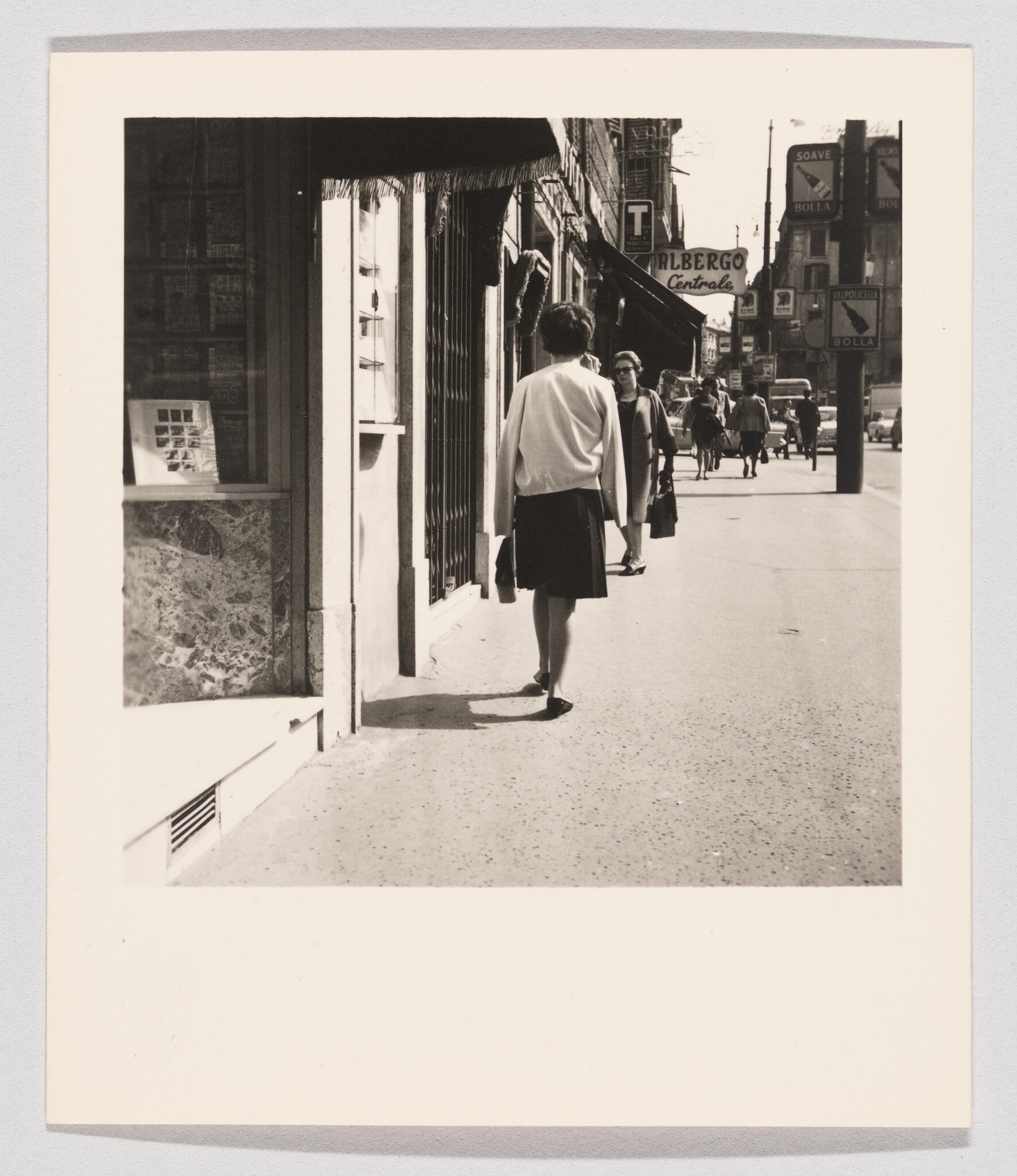 A woman in a skirt walks down a busy city sidewalk carrying a handbag.