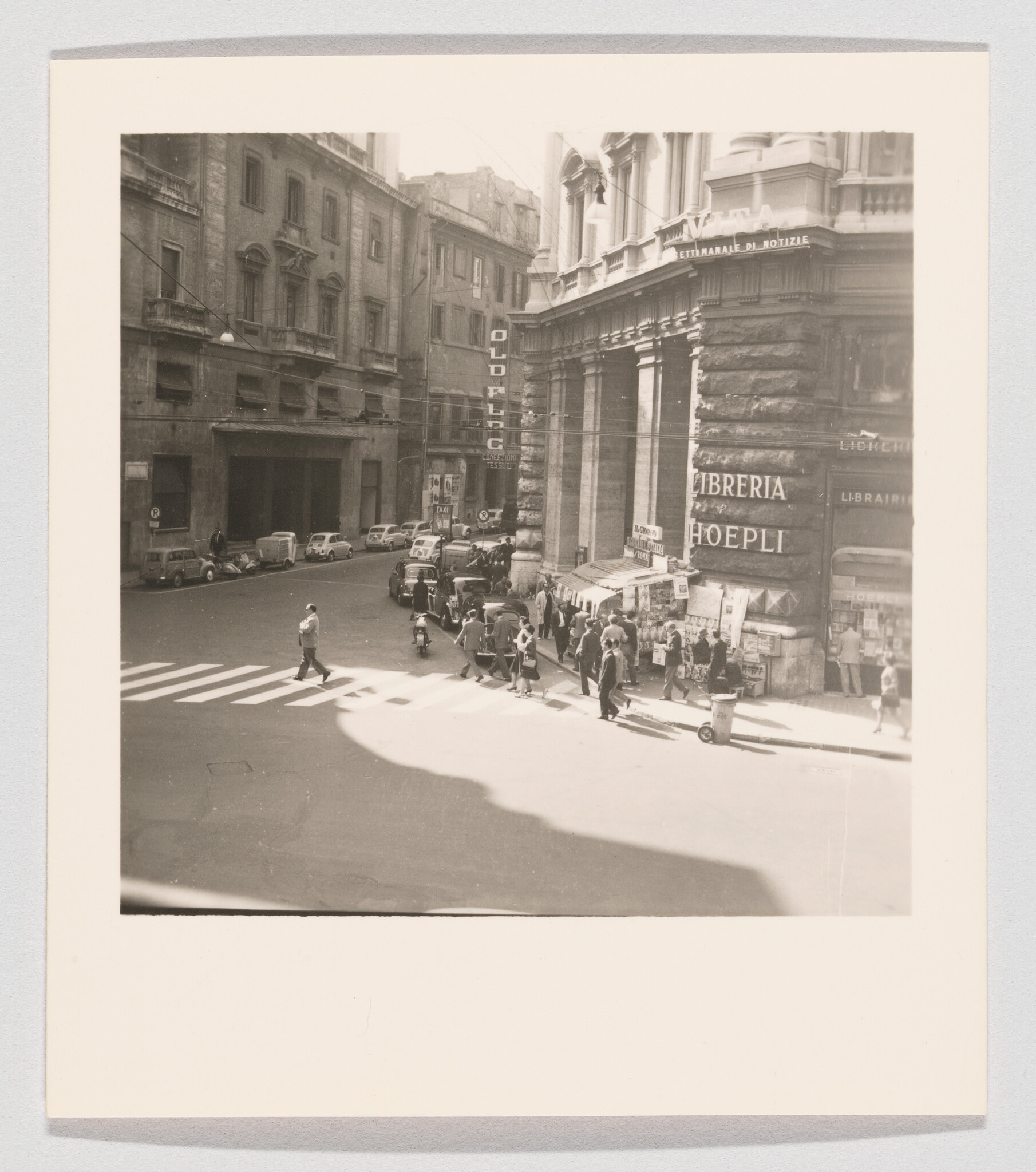 Pedestrians cross a busy street corner outside the Libreria Hoepli bookstore with vintage cars parked.