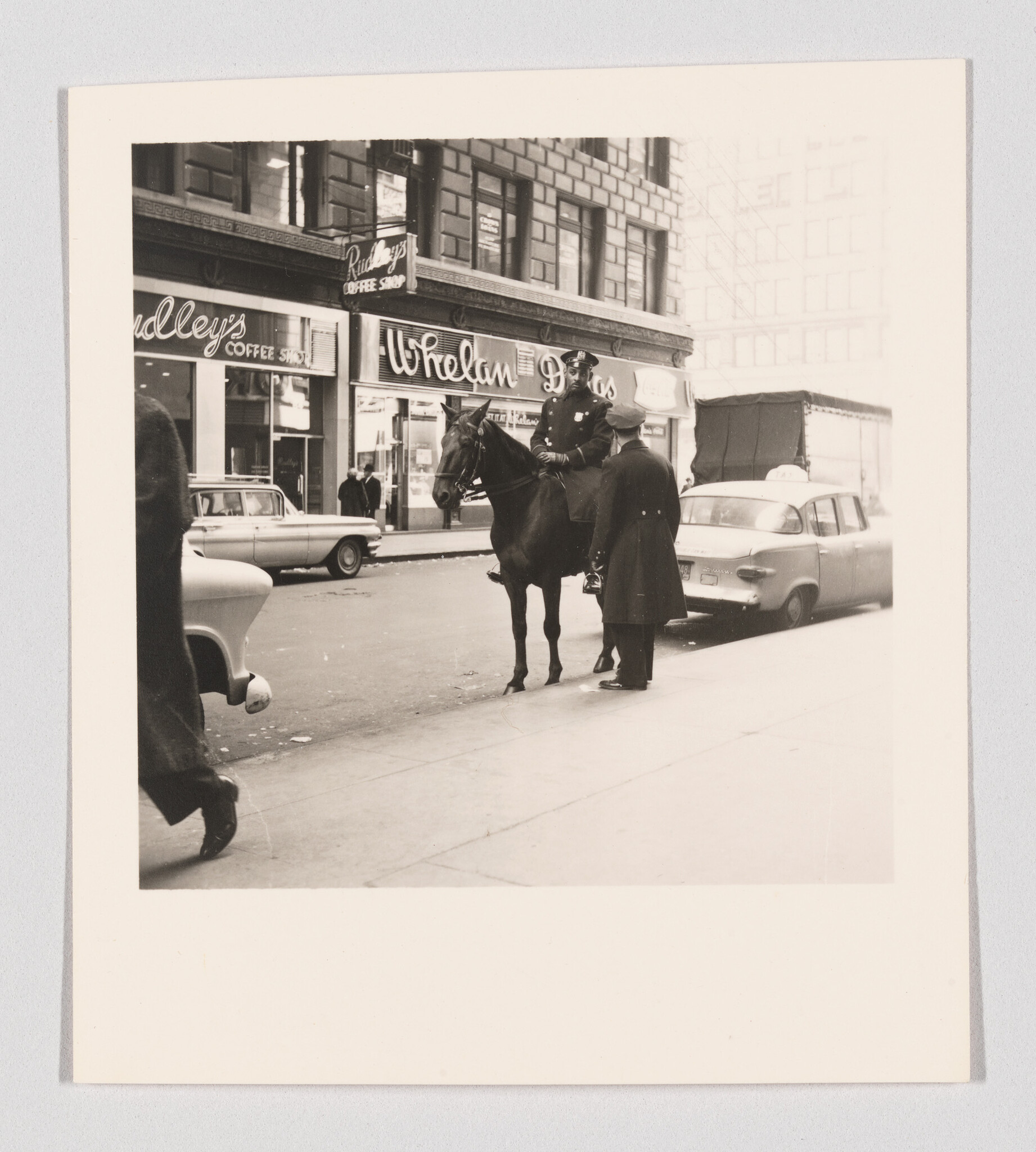 A vintage black and white photograph depicting an urban street scene with a mounted police officer on a horse talking to two individuals on a sidewalk. In the background, there are storefronts with neon signs, including a coffee shop, and a classic car parked on the street.