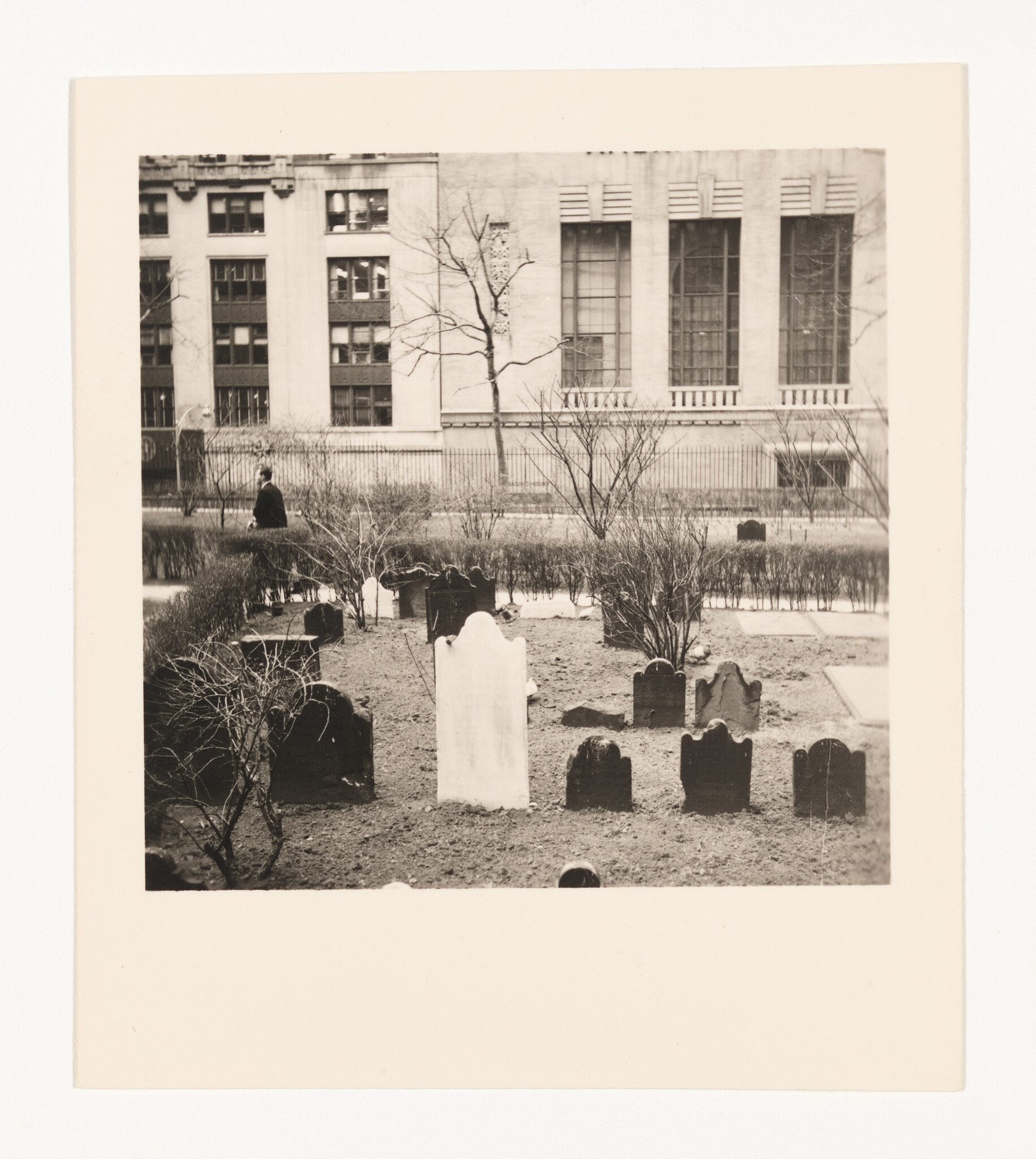 Small historic graveyard with worn headstones and a prominent white tombstone in front of a city building.
