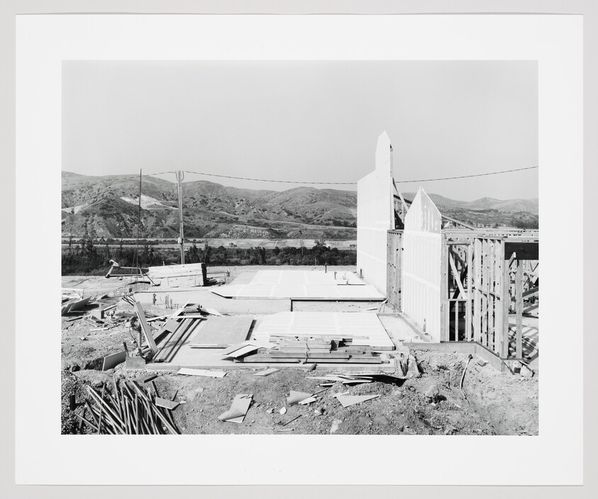 House under construction with foundation and framed walls amid hilly landscape.