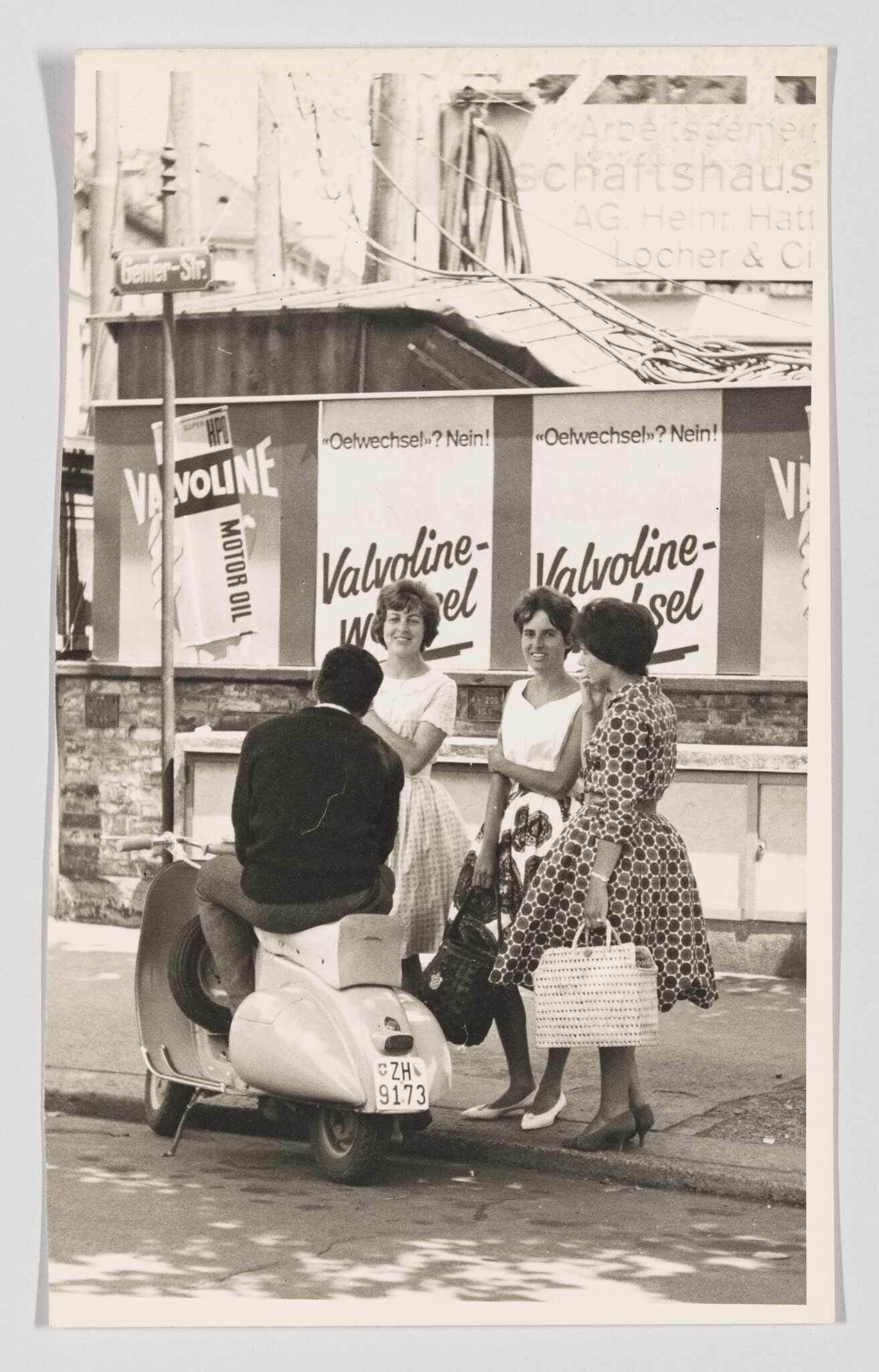 Three women chat with a man sitting on a scooter in front of Valvoline advertisement.