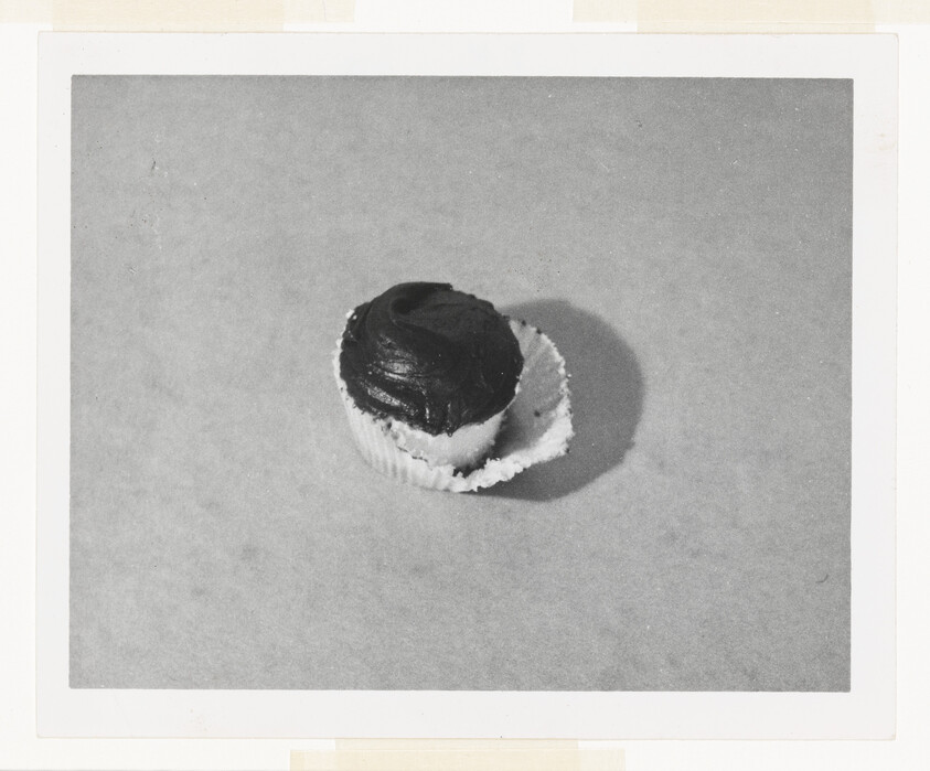 A chocolate-frosted cupcake with its paper liner partially peeled away on a plain surface.