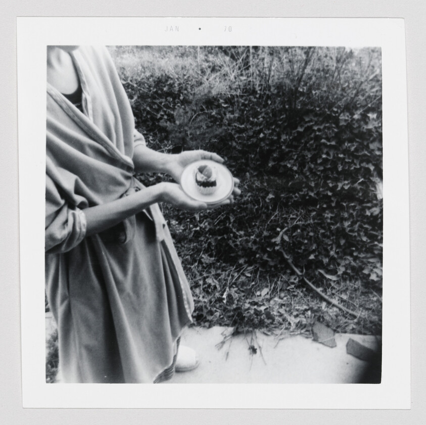 Person holding a small plate with a cupcake while standing by overgrown ivy.