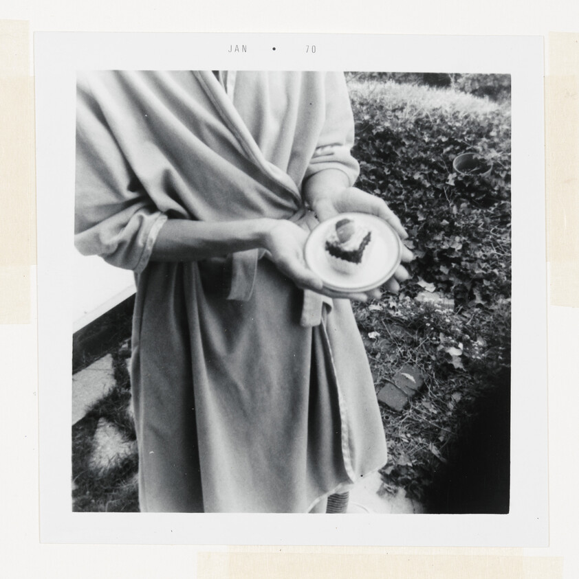 Person in a robe holds a small plate with a single slice of cake near a garden.