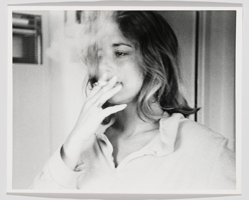 A young woman stands indoors smoking a cigarette as smoke drifts around her face.