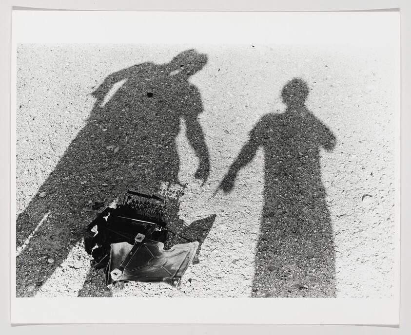 Two people's shadows fall over a broken typewriter on a gravel surface.
