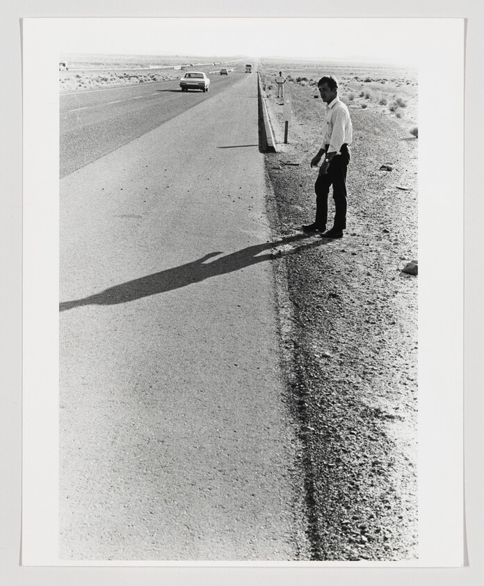 A man stands at the side of a desert highway casting a long shadow.