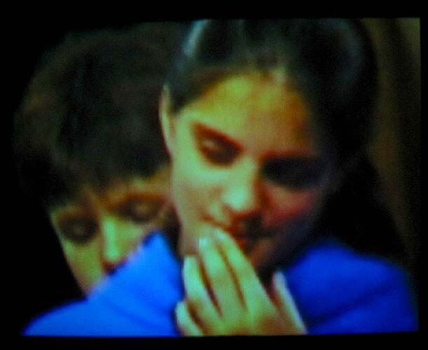 A young woman in a blue sweater looks down and touches her mouth while another person stands behind her.