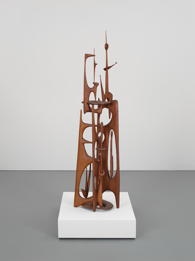 Abstract wooden sculpture with intricate cutouts and tall, thin shapes on a white pedestal.