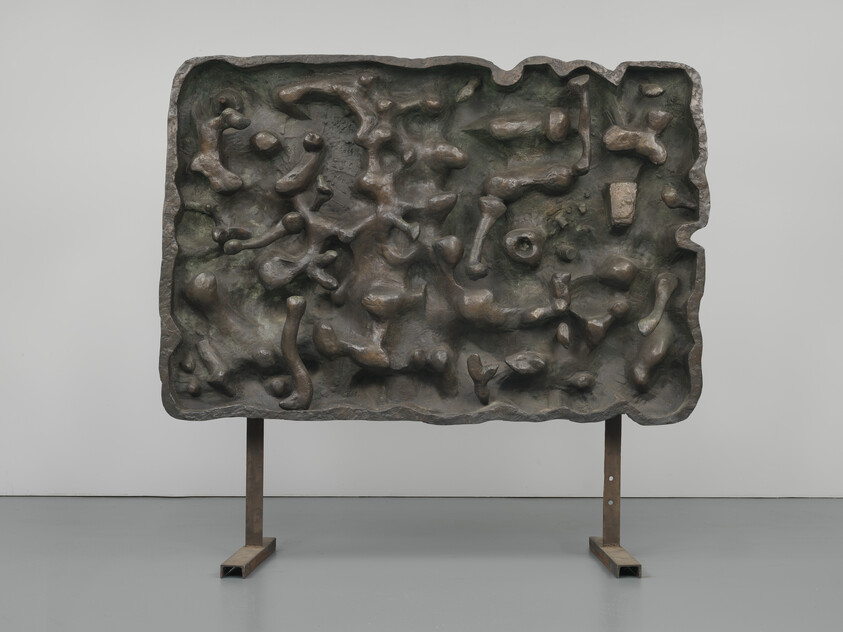 Large abstract bronze relief with organic bulges mounted on a simple metal stand in a gallery.