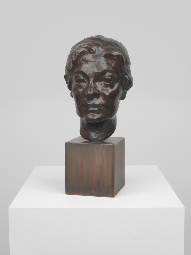 Bronze sculpted head of a woman displayed on a square bronze base atop a white plinth.