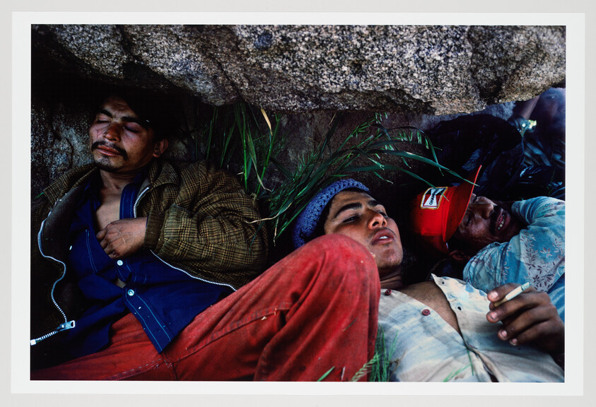 Three men lie resting under a rock ledge, one holding a cigarette.