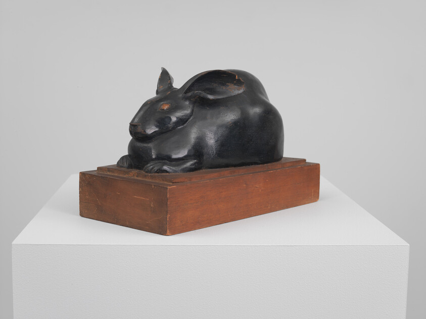 A small black rabbit sculpture rests on a rectangular wooden base on a white pedestal.
