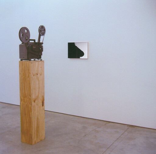 Old film projector sits on a wooden pedestal in a minimalist gallery next to a small black artwork.