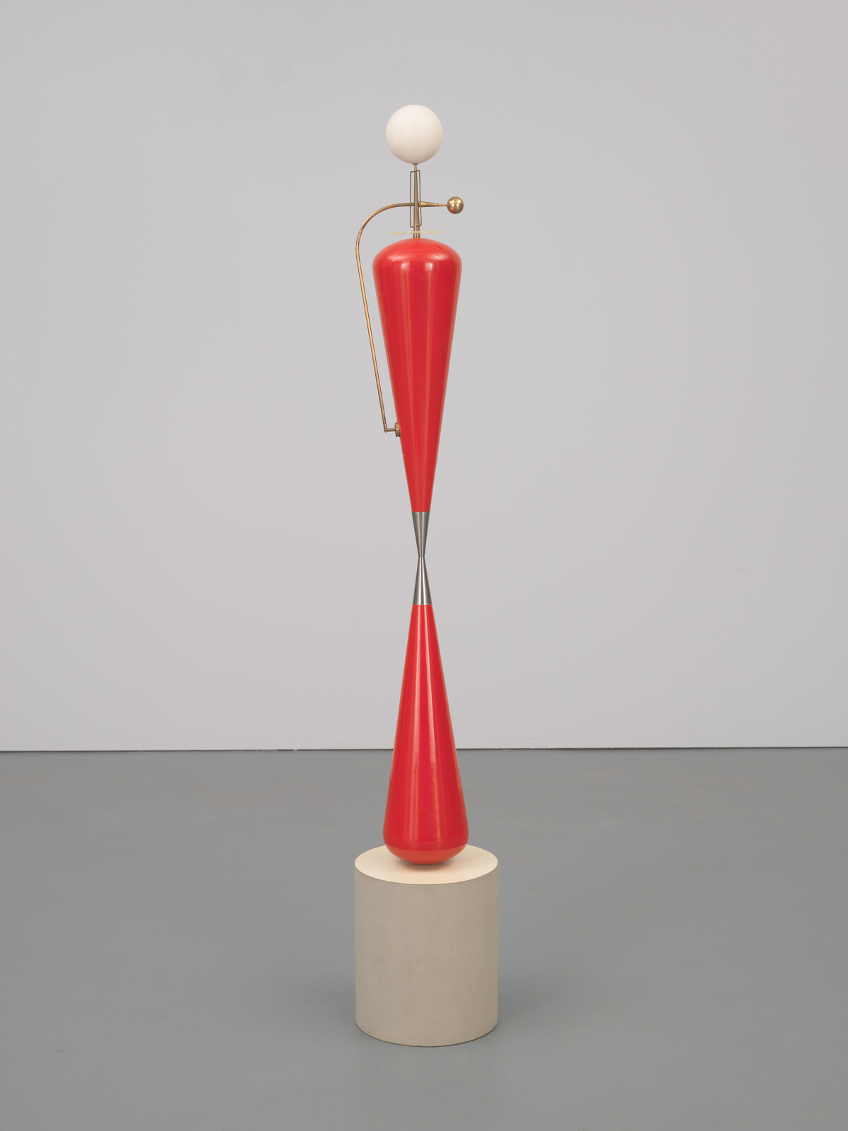 A tall red double-cone sculpture with a white ball and brass arm stands on a round pedestal.