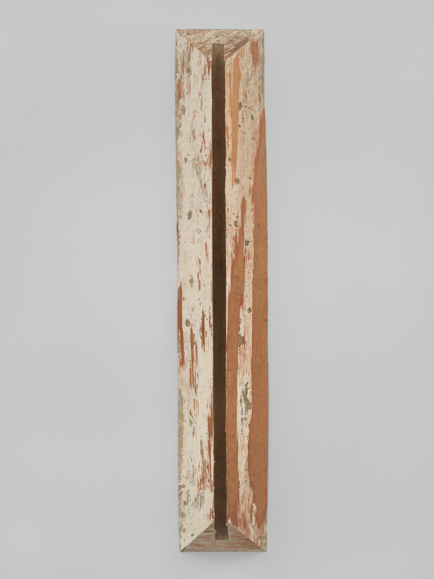A long, narrow wooden sculpture with a vertical slit down the middle, showing signs of weathering and peeling white paint, displayed against a plain white background.