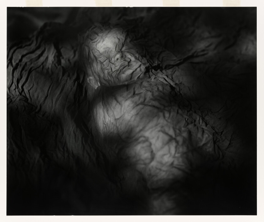 A shrouded figure lies under crumpled cloth with light revealing facial and chest contours.