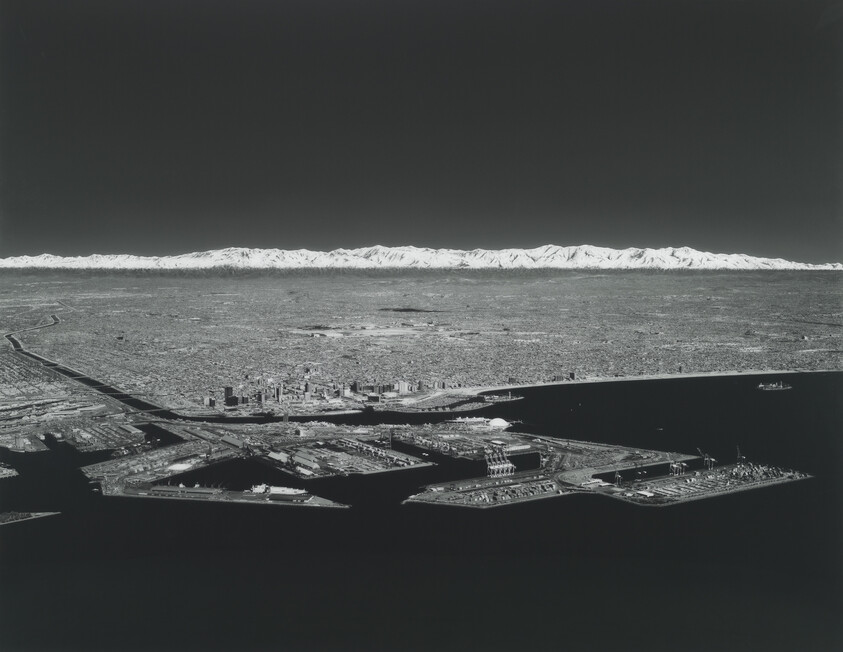 Aerial view of a busy coastal port and city with snow-capped mountains on the horizon.