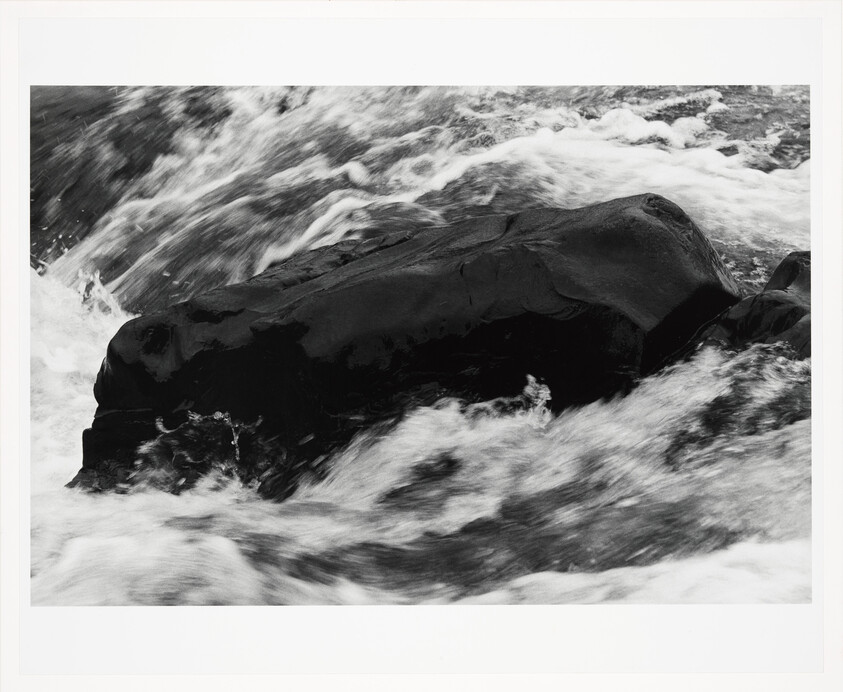 A dark boulder stands in fast moving whitewater as waves crash around it.