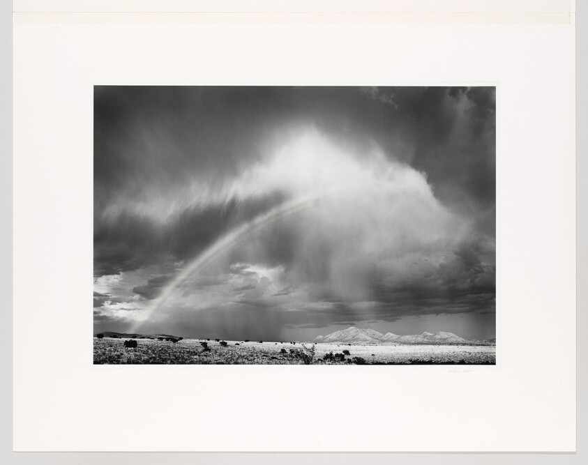 A wide plain with a faint rainbow arcing through heavy rain clouds over distant mountains.