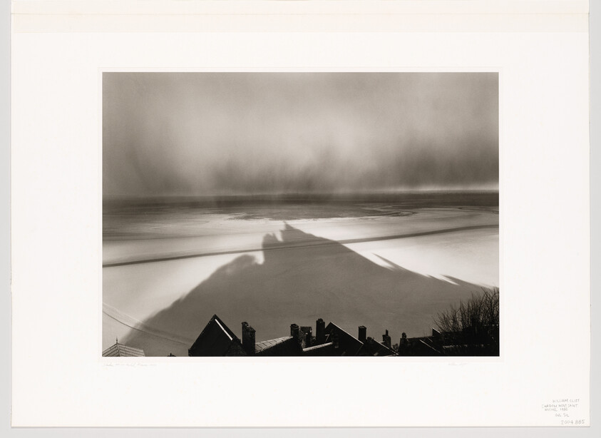 Rooftops in the foreground with a large mountain shadow stretching across a snowy plain under a misty sky.