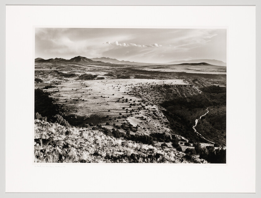 Wide black-and-white view of a sunlit plateau and winding canyon with distant mountains.