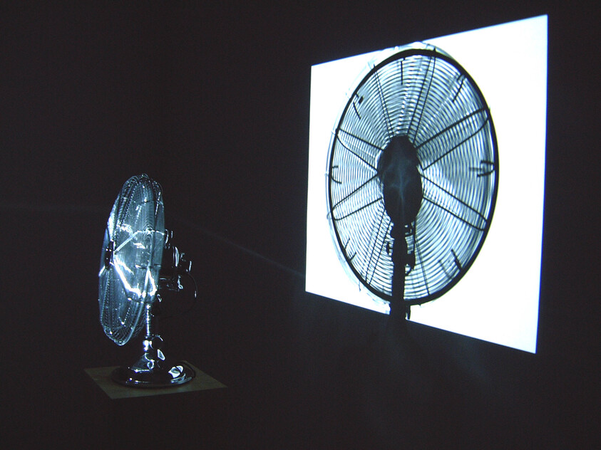 A desktop oscillating fan projects a large shadow onto a bright white screen.